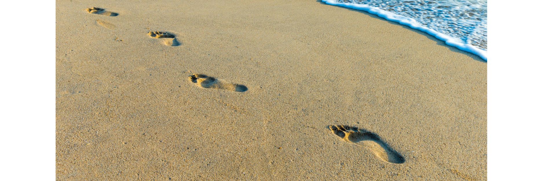 Four bare human footprints pressed into smooth beach sand, leading from the lower right toward the upper right where a thin wave is washing up onto the shore.