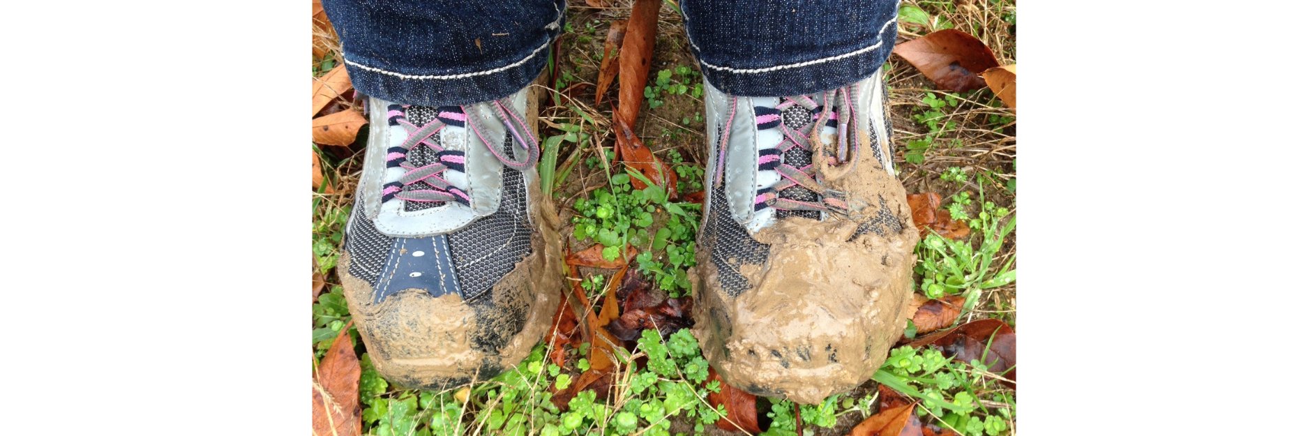 A pair of athletic shoes with pink and black laces, photographed from above. The toes of both shoes are thickly caked in wet brown mud. The shoes are standing on grass dotted with small green clover-like plants and scattered fallen brown leaves. The cuffs of dark blue jeans are visible at the top.