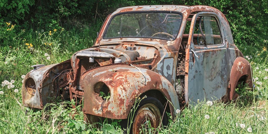 An old car whose metal is covered in an orange-brown flaky coating, with empty window openings, standing in tall grass and weeds.